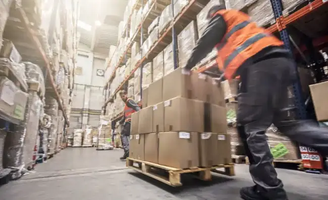 Warehouse workers in hi-vis vests moving boxes on a pallet among tall storage shelves